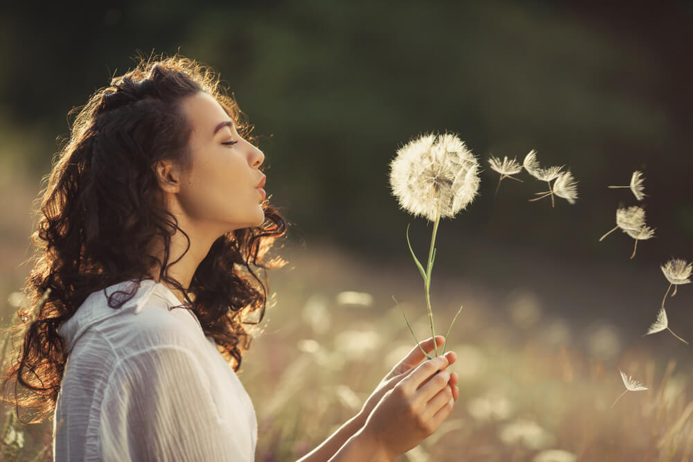 Woman blowing dandelion seeds in a sunlit field, symbolizing letting go and healing